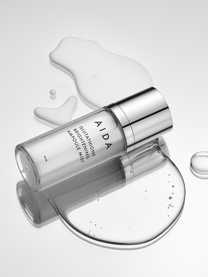 an AIDA Glutathione Brightening Ampoule Mist bottle lying on a reflective surface with its contents artistically spilled, highlighting the clear and sleek design of the bottle and the purity of the skincare product