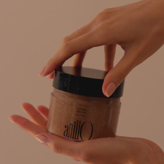  hand gently opens a jar of Fig Whisky Body Scrub by anillo, revealing its textured contents, ready for use. The focus is on the natural ingredients visibly incorporated into the scrub, poised to offer a rejuvenating skincare experience.